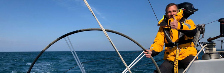 Zdjęcie na pokładzie żaglówki przedstawiające osobę w pomarańczowej kurtce żeglarskiej trzymającą za ster i patrzącą w obiektyw z kciukiem do góry
